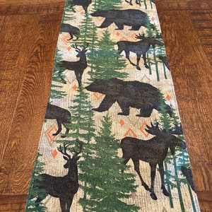 May include: A beige table runner with a woodland theme. The runner features a repeating pattern of black bears, moose, deer, and pine trees on a beige background with orange diamond shapes.