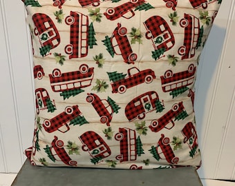 May include: A red and black plaid pillow with a white background featuring a repeating pattern of red trucks carrying Christmas trees. The trucks are decorated with green holly and red berries.