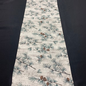 May include: A rectangular table runner with a repeating pattern of pine branches and pine cones. The branches are green and brown, and the cones are brown. The background is a light beige color with a wood grain texture.