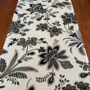 May include: A white table runner with a black and gray floral pattern. The pattern features paisley-like designs and intricate details.