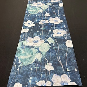 May include: A blue table runner with a white floral pattern. The flowers are stylized and have a watercolor effect. The runner is made of a soft, textured fabric.