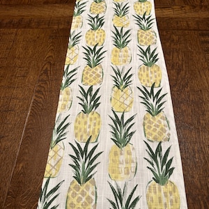 May include: A white table runner with a pineapple pattern. The pineapples are yellow and green and are arranged in rows. The runner is made of a linen-like fabric.