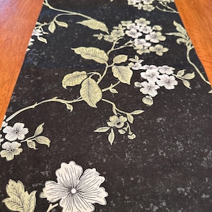 May include: Black table runner with a floral pattern in shades of green and white. The flowers are stylized and have a vintage look.