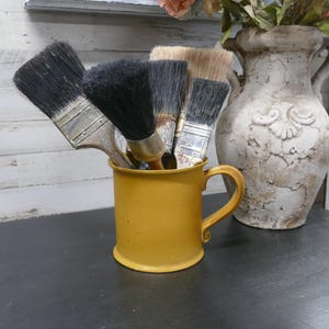 Puede incluir: Un conjunto de pinceles con cerdas negras en una taza de color amarillo mostaza con asa. La taza está sobre una superficie oscura, con un jarrón decorativo y flores en el fondo.