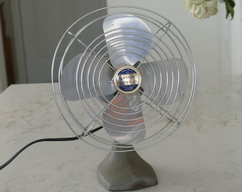 May include: A vintage Manning Bowman electric desk fan with a silver-toned metal cage and four metallic blades. The center of the fan features a circular blue and gold emblem with the text Manning Bowman. It has a heavy gray base and a black cord.