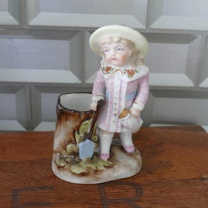 May include: A vintage porcelain figurine of a young girl in a pink coat and white hat, holding a shovel and a watering can. The figurine is standing next to a tree stump-shaped planter. The planter is brown with green leaves and a blue shovel.