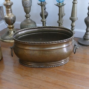 Peut inclure: Un grand bol en métal de couleur bronze antique avec une bordure en forme de corde sur le dessus et le bas. Le bol a deux petites poignées et repose sur un sol en bois. Plusieurs chandeliers décoratifs en laiton sont en arrière-plan.