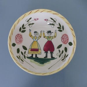 Puede incluir: Un cuenco redondo de cerámica con borde amarillo y diseño pintado. El cuenco presenta dos figuras con vestimenta tradicional, rosas rosas y follaje verde. Las figuras levantan los brazos en señal de celebración.