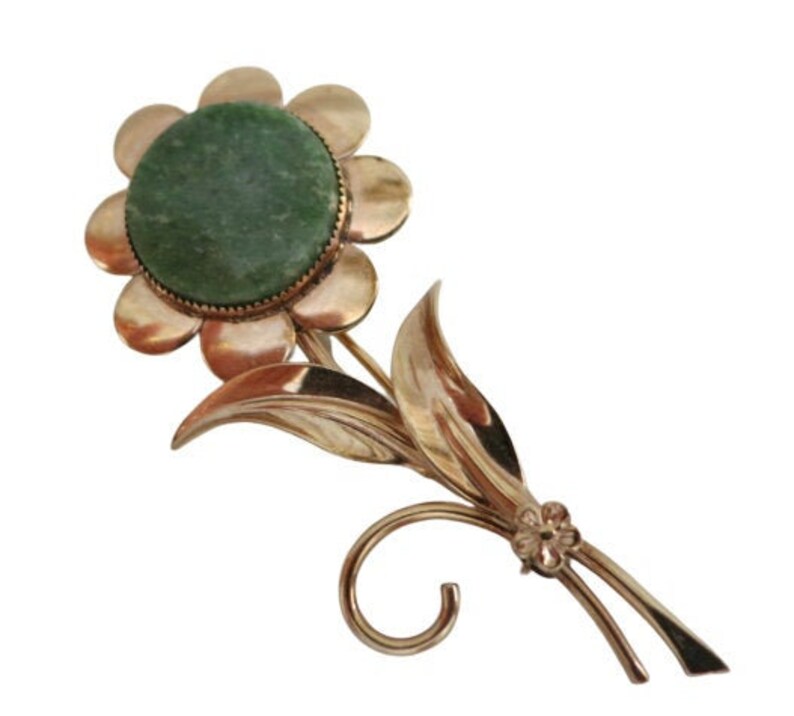 May include: A gold-toned brooch in the shape of a flower with a green stone in the center. The flower has a round center and eight petals. The stem has two leaves and a small flower at the end.