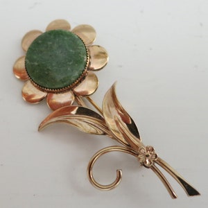 May include: A gold-toned brooch in the shape of a flower with a green stone center. The flower has a round center and eight petals. The stem has two leaves and a small flower at the bottom.