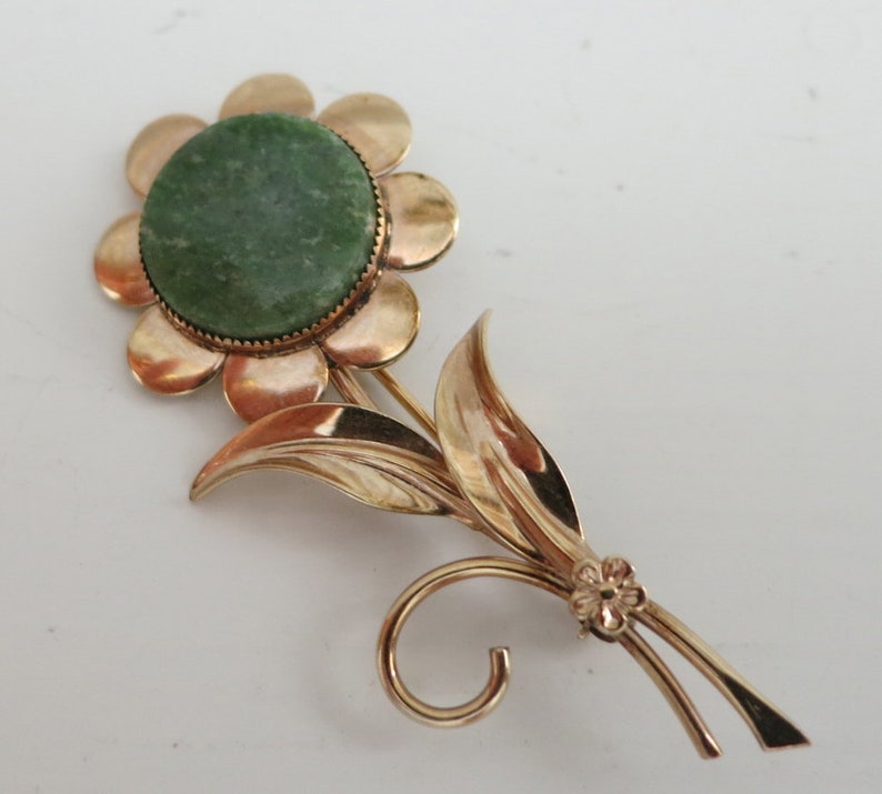 May include: A gold-toned brooch in the shape of a flower with a green stone center. The flower has a round center and eight petals. The stem has two leaves and a small flower at the bottom.