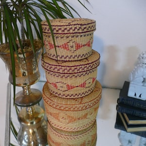 May include: Three woven storage baskets stacked on top of each other. The baskets are round with lids and feature a repeating pattern of red and purple geometric designs. The baskets are made of a natural fiber material.