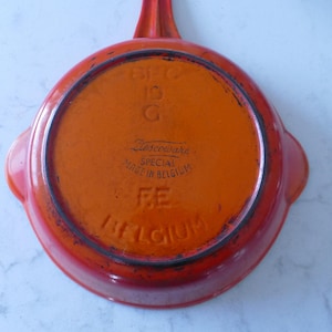 Könnte beinhalten: Eine orangefarbene gusseiserne Pfanne mit Griff und zwei Seitengriffen. Auf der Unterseite der Pfanne steht "Ziescoware SPECIAL MADE IN BELGIUM FE BELGIUM". Die Pfanne ist rund und hat einen dunkelorangen Innenraum.