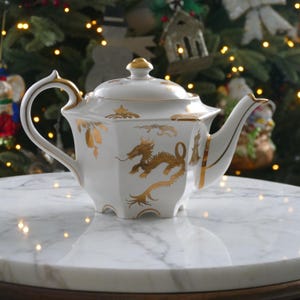 May include: White porcelain teapot with gold dragon and floral designs. The teapot has a lid with a gold knob, a curved handle, and a spout. The teapot is sitting on a marble table.