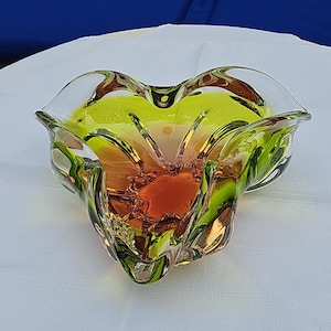 May include: A decorative glass bowl featuring a vibrant gradient of lime green and amber orange. The bowl has a distinct ruffled, organic rim with clear glass edges and a thick, translucent body that catches the light, creating depth.