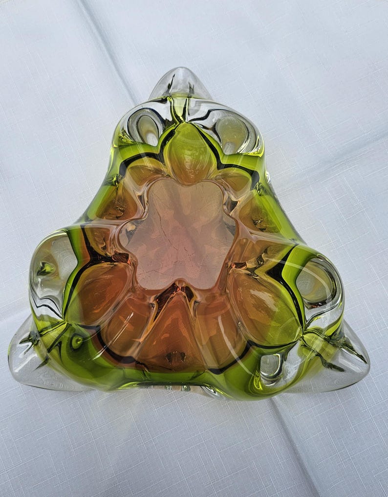 May include: A triangular glass bowl featuring a vibrant gradient of lime green and amber orange. The thick, clear glass edges form a scalloped, multi-lobed interior design with a deep central basin, creating a complex, layered visual effect.