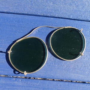 Puede incluir: Gafas de sol vintage con clip, con lentes verde oscuro y montura de metal plateado. Las lentes redondas están conectadas por un puente curvo. Las gafas de sol están sobre una superficie de madera azul.