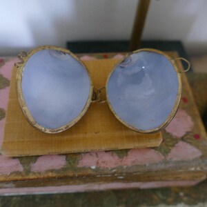 May include: Vintage spectacles with light blue lenses and gold-coloured frames. The lenses are oval-shaped and set in a delicate, ornate frame. The spectacles are open, revealing the light blue lenses. The spectacles are resting on a book.