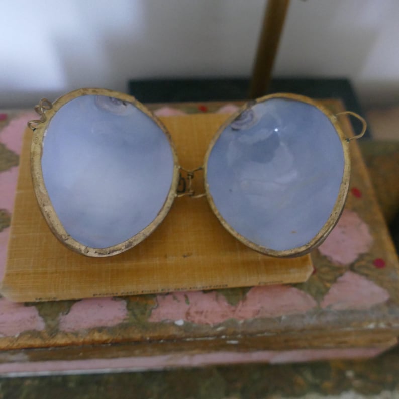 May include: Vintage spectacles with light blue lenses and gold-coloured frames. The lenses are oval-shaped and set in a delicate, ornate frame. The spectacles are open, revealing the light blue lenses. The spectacles are resting on a book.