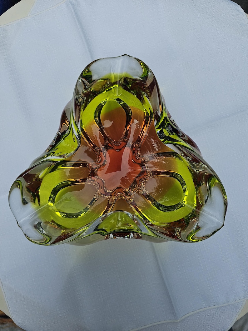 May include: A thick-walled glass bowl with a three-lobed, triangular shape. The glass features a vibrant gradient transition from a deep amber-orange center to a bright lime-green rim, with clear glass edges defining the sculptural form.