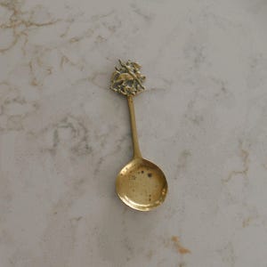 May include: A brass spoon featuring a decorative handle top with a sculpted relief of a winged figure. The bowl of the spoon has a smooth, polished surface with visible dark patina spots, and the handle is a simple, slender rod connecting to the top.