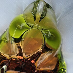 May include: A sculptural glass bowl featuring a vibrant lime green rim that transitions into a deep amber and brown center. The piece has an organic, asymmetrical shape with thick, clear glass walls and smooth, flowing curves throughout the design.