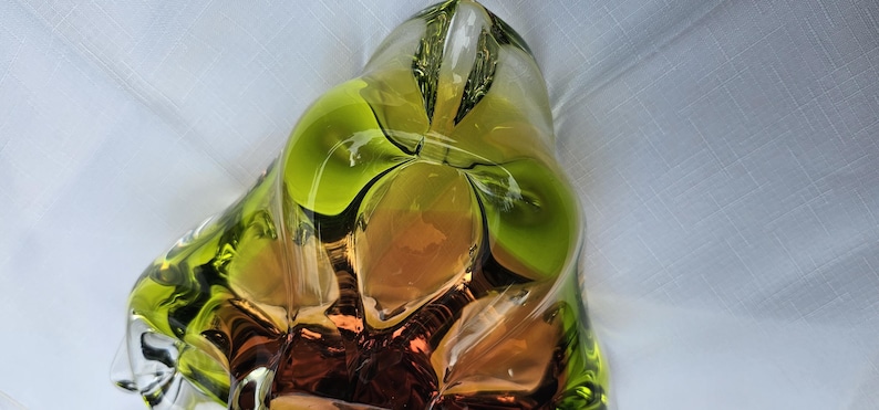 May include: A sculptural glass bowl featuring a vibrant lime green rim that transitions into a deep amber and brown center. The piece has an organic, asymmetrical shape with thick, clear glass walls and smooth, flowing curves throughout the design.
