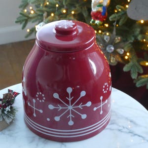 Op de afbeelding: Een rode keramische koekjespot met een deksel, versierd met witte sneeuwvlokontwerpen. De pot heeft een afgeronde vorm en is versierd met witte strepen aan de basis. Een kleine sneeuwvlok siert het deksel.
