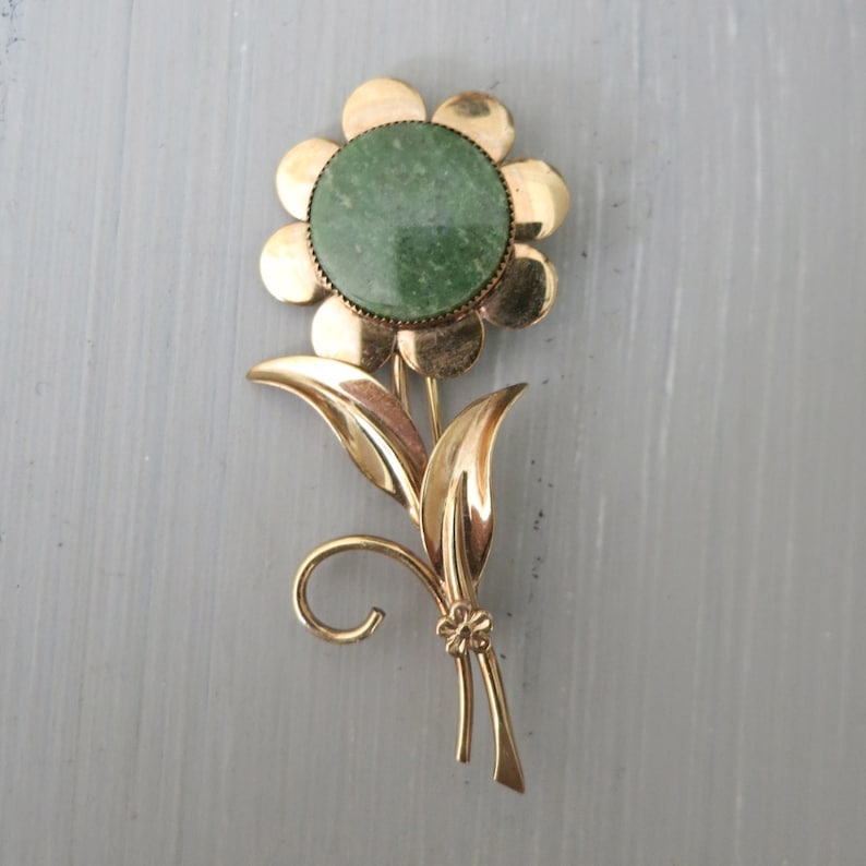 May include: A gold-tone flower brooch with a green stone center. The flower has a simple design with five petals and a stem with two leaves.