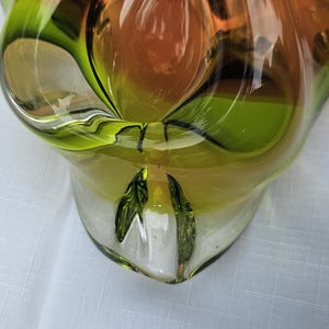 May include: A decorative glass bowl featuring a vibrant blend of translucent lime green and warm amber tones. The piece has a smooth, polished surface with organic, undulating curves and a thick, clear glass base that highlights the color gradient.