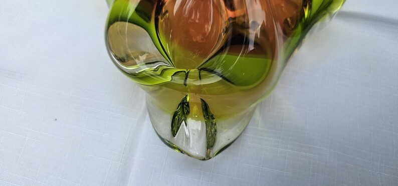 May include: A decorative glass bowl featuring a vibrant blend of translucent lime green and warm amber tones. The piece has a smooth, polished surface with organic, undulating curves and a thick, clear glass base that highlights the color gradient.