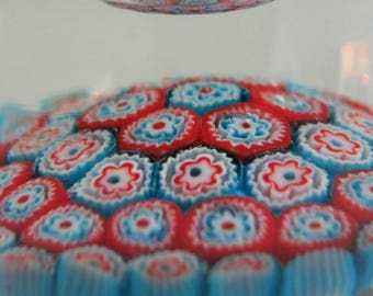 May include: A glass paperweight with a colorful, multi-layered design. The paperweight features a pattern of red, white, and blue circles, resembling a flower.
