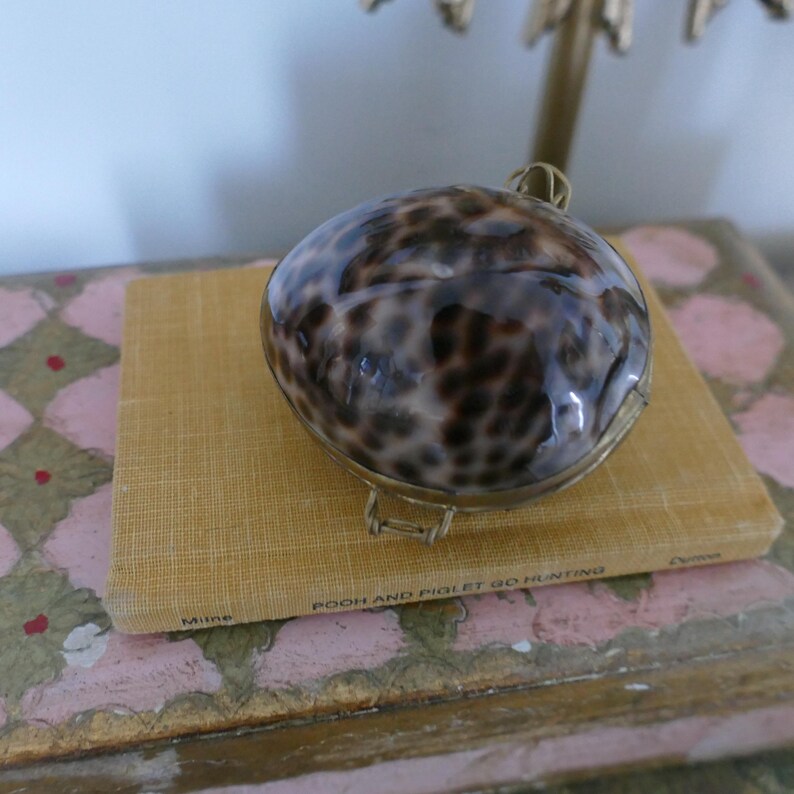 May include: A decorative, round trinket box with a shell-like pattern in shades of brown and tan. The box has a gold-colored metal frame and is resting on a small, rectangular book with the title "POOH AND PIGLET GO HUNTING".
