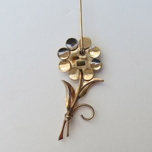 May include: Gold tone flower brooch with a pin back. The flower has a circular design with multiple round petals. The stem has two leaves and a curled end.