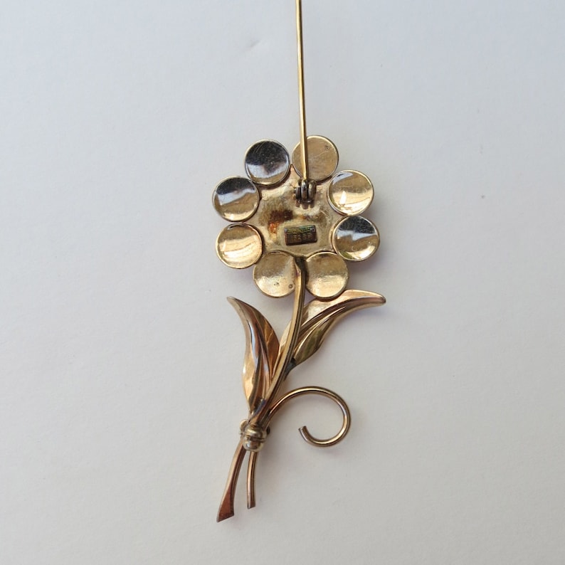 May include: Gold tone flower brooch with a pin back. The flower has a circular design with multiple round petals. The stem has two leaves and a curled end.