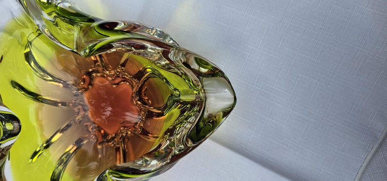 May include: A decorative glass bowl featuring a vibrant lime green outer rim that transitions into a warm amber orange center. The bowl has a distinct organic, undulating shape with thick, clear glass edges that create a layered, translucent effect.