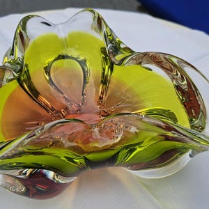 May include: A decorative glass bowl featuring a vibrant chartreuse green rim that transitions into a warm amber center. The bowl has a distinct organic, free-form shape with undulating, ruffled edges and a smooth, polished finish throughout.