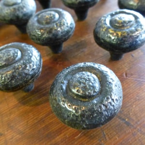 May include: Close-up of several dark gray, textured cabinet knobs with a hammered metal appearance. Each knob has a circular design with a raised center and a short stem. The knobs are arranged on a wooden surface.