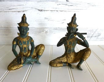 May include: Two bronze-colored statues of seated figures in traditional attire. One figure holds a flute to their lips, while the other holds an object in their hands. The statues have gold-colored accents on their clothing and headwear.