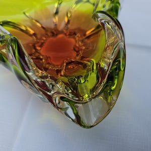 May include: A decorative glass bowl featuring a vibrant gradient of colors, transitioning from a deep amber orange at the center to a bright lime green along the scalloped, undulating edges. The glass has a smooth, polished finish with clear sections.