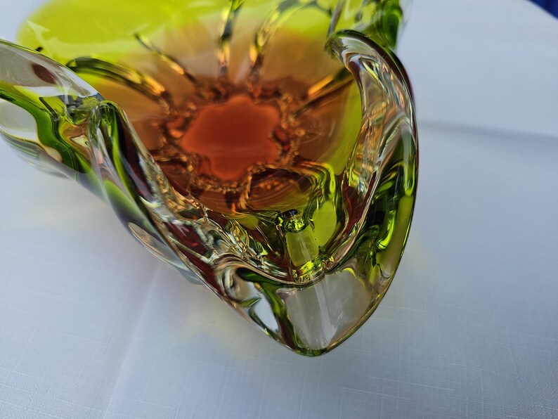 May include: A decorative glass bowl featuring a vibrant gradient of colors, transitioning from a deep amber orange at the center to a bright lime green along the scalloped, undulating edges. The glass has a smooth, polished finish with clear sections.