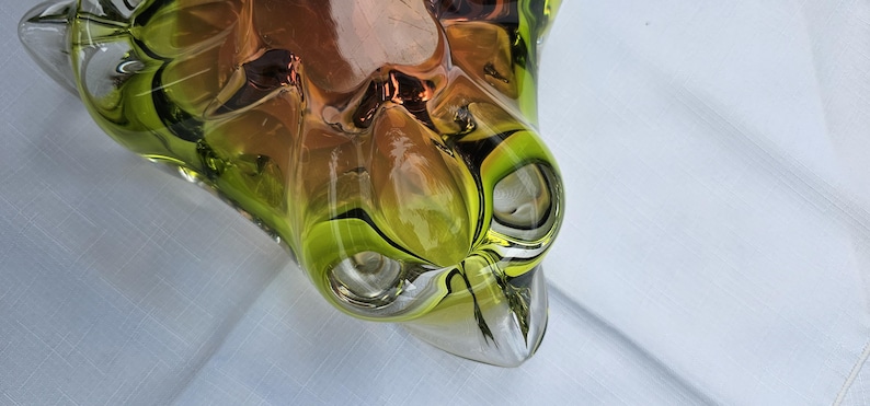 May include: A decorative glass bowl featuring a vibrant gradient of lime green and amber tones. The bowl has a thick, clear glass rim and a complex, multi-faceted organic shape with deep indentations and smooth, rounded protrusions throughout.