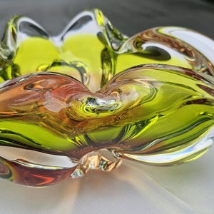 May include: A decorative glass bowl featuring a vibrant chartreuse green center that transitions into clear glass edges. The bowl has a distinct ruffled, undulating rim with a fluid, organic shape and a smooth, polished finish throughout the piece.