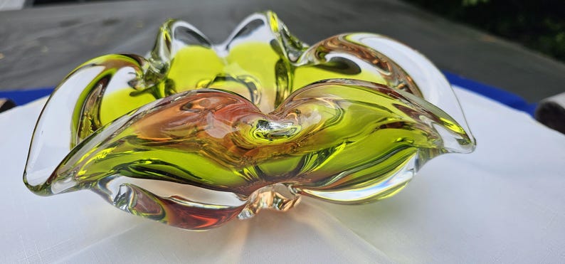 May include: A decorative glass bowl featuring a vibrant chartreuse green center that transitions into clear glass edges. The bowl has a distinct ruffled, undulating rim with a fluid, organic shape and a smooth, polished finish throughout the piece.
