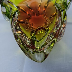 May include: A decorative glass bowl featuring a vibrant gradient of lime green and deep orange. The bowl has a thick, clear glass rim with a wavy, organic edge and a smooth, polished surface that reflects light, creating a multi-dimensional appearance.