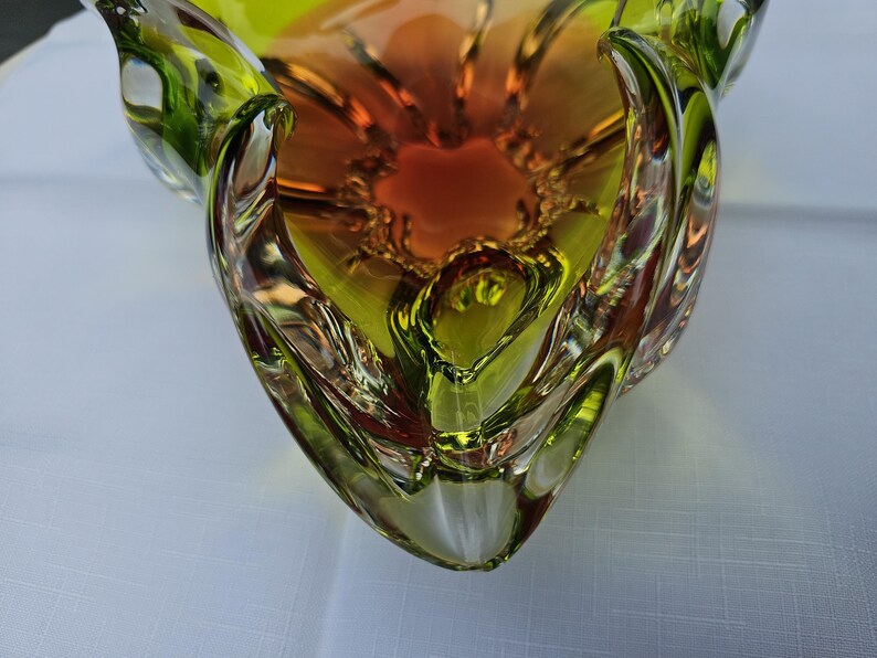 May include: A decorative glass bowl featuring a vibrant gradient of lime green and deep orange. The bowl has a thick, clear glass rim with a wavy, organic edge and a smooth, polished surface that reflects light, creating a multi-dimensional appearance.