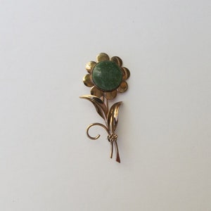 May include: A gold-tone flower brooch with a green stone center. The flower has a simple design with rounded petals and a stem with leaves.