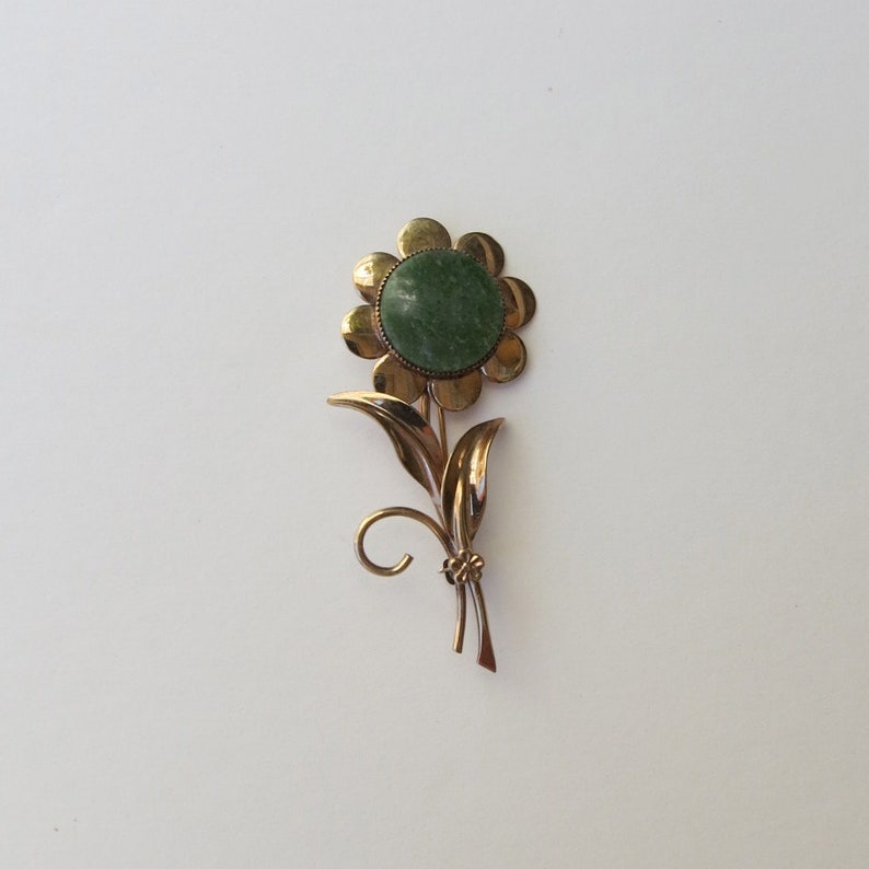 May include: A gold-tone flower brooch with a green stone center. The flower has a simple design with rounded petals and a stem with leaves.