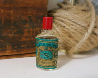 Puede incluir: Una pequeña botella de vidrio transparente de Eau de Cologne con tapa roja. La etiqueta presenta detalles en turquesa y dorado con el texto "4711". La botella es probablemente vintage y está cerca de una caja de madera y una madeja de cuerda.