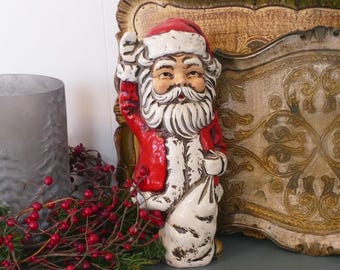 May include: A vintage Santa Claus figurine, painted in red, white, and beige. The figurine is holding a sack and has a long white beard. The Santa Claus figure is a festive holiday decoration.